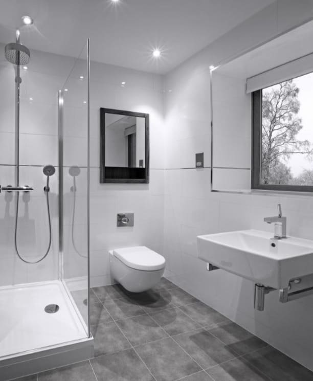All white bathroom, shower on the left of the toilet, there is a mirror above the toilet and sink in front of it with a window above the sink, grey floor